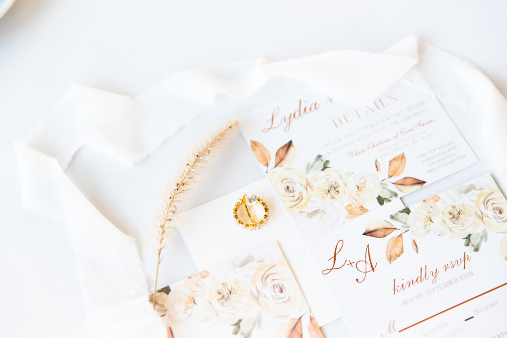 Soft boho floral wedding invitation with a white ribbon and gold engagement ring and wedding band with postage stamps.